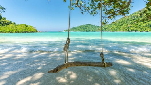 A swing on a beach in Thailand with an idyllic view of the water and lush hills.