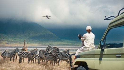 A safari traveller sat on a jeep observes zebras in the wild in front of a lake and mountains