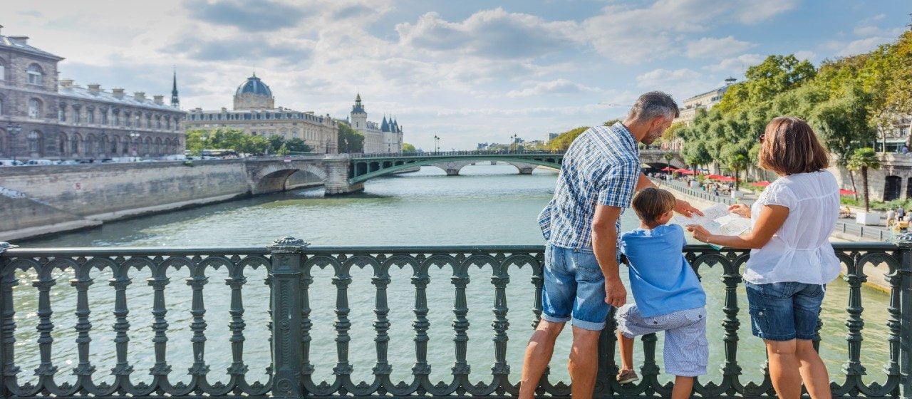 A family of three looking at a map on a bridge over the river in central Paris on a sun-soaked day