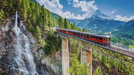 Train in Switzerland on a bridge with mountains in the background and trees below on a beautiful day.